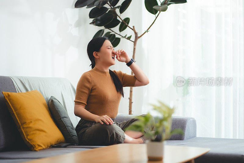 亚洲妇女发烧，流鼻涕，咳嗽和打喷嚏，坐在家里的沙发上。