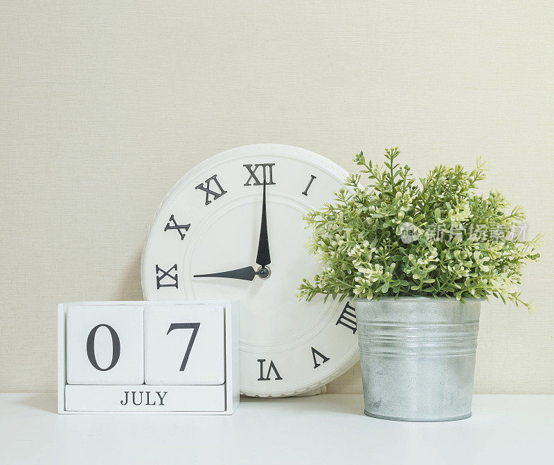 白色木历以黑色7月7日字与时钟和植物搭配白色木桌和奶油色壁纸纹理背景，选择性聚焦在日历上