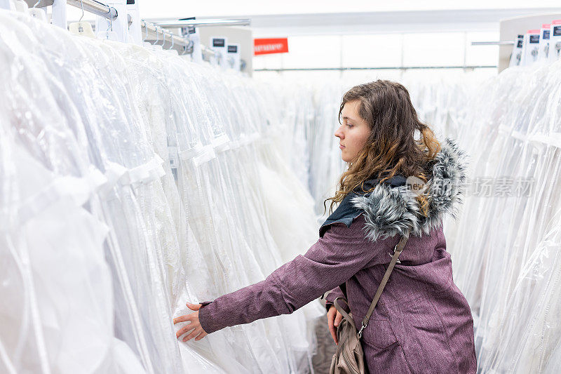 年轻女子在精品折扣店寻找触摸一件蕾丝婚纱，许多白色服装挂在衣架排