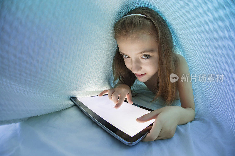 小女孩睡在羽绒被下使用她的电子平板电脑