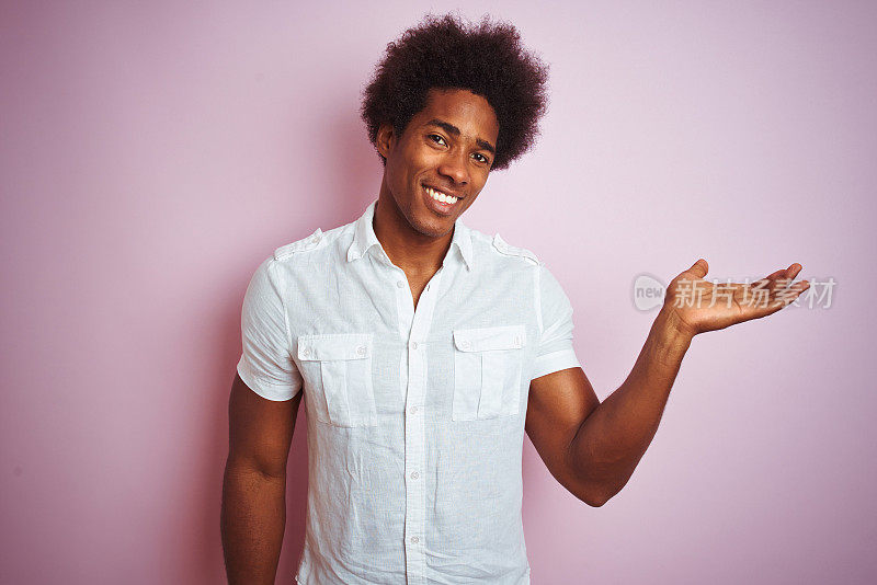 一个年轻的美国男子，留着非洲式发型，穿着白衬衫，站在孤立的粉红色背景上，面带微笑，用手掌指着镜头。