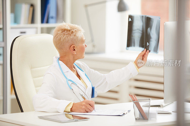 合格的成熟女性骨科医师穿着白大褂坐在桌子旁，检查膝盖的x光片