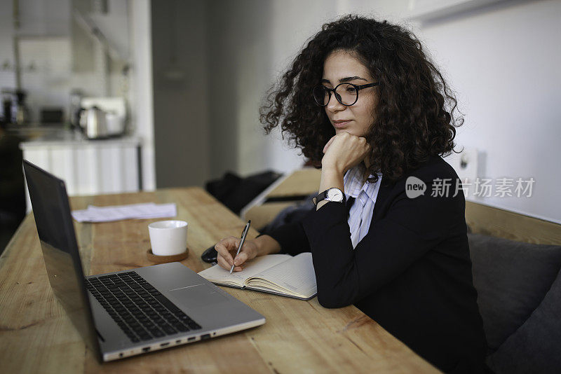 女商人在办公室看着笔记本电脑做笔记。女企业家坐在桌子边写笔记边用笔记本电脑工作。