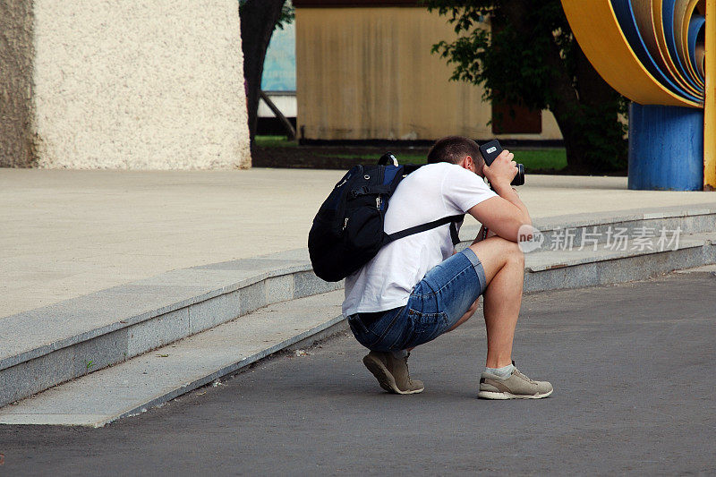 一名男子身穿牛仔裤短裤、白色t恤、黑色背包，手里拿着相机和智能手机，蹲下来为某人或户外旅游景点拍照