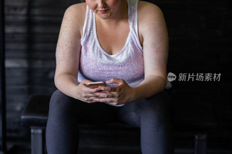 锻炼休息:一个超重的女人在健身房使用她的手机