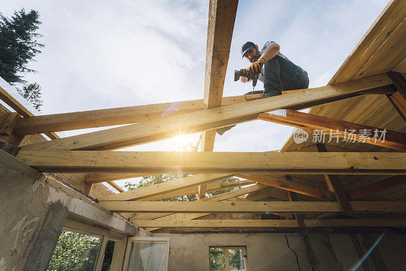 背光工人在不完整的住宅结构屋顶上使用电钻