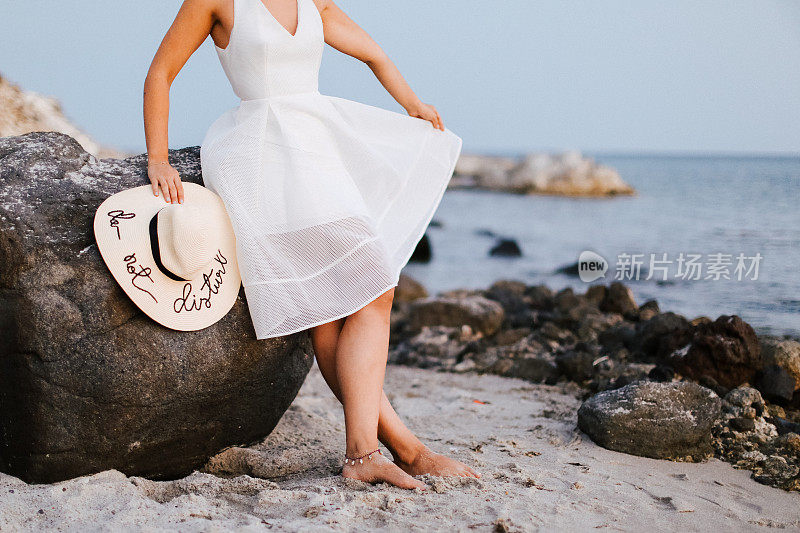 美丽的女人戴着草帽，穿着白色的衣服走在海边的海滩上