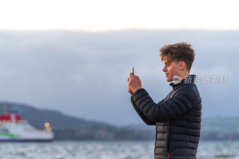 一个十几岁的男孩在海滩上用手机拍照