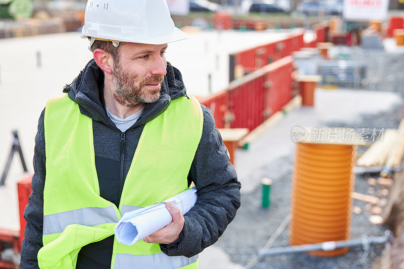 一名建筑师或土木工程师戴着白色安全帽，拿着卷起来的规划文件和一件安全背心站在建筑工地上正在施工的地基前。