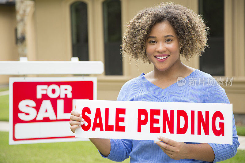 可爱的年轻成人房地产经纪人站在她旁边的出售标志在家门口的院子里。她拿着一个待售的牌子，打算放在招牌的顶端。