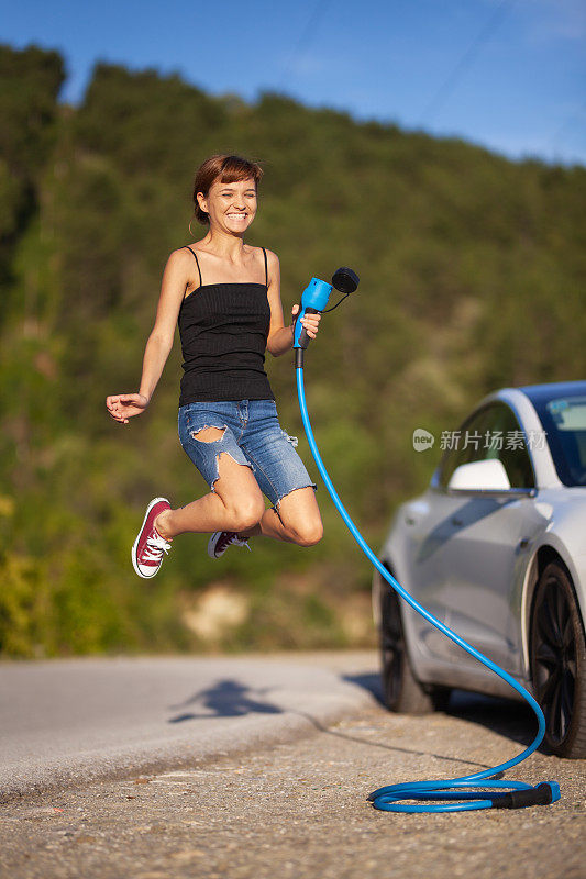 一个年轻漂亮的女孩跳到一辆电动汽车旁边。拿着蓝色充电线