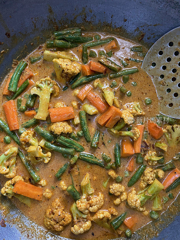 花椰菜、切碎的胡萝卜和青豆在厨房灶具上的不粘锅中烹饪的特写图像，用咖喱酱油煨蔬菜，用有槽的金属勺搅拌