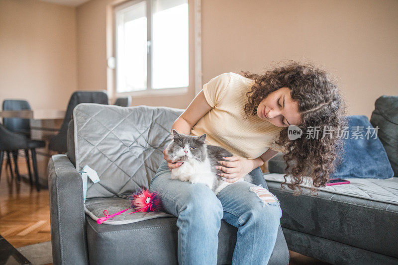 卷发的年轻女子坐在沙发上，和她那只灰白色的猫和它的玩具玩耍
