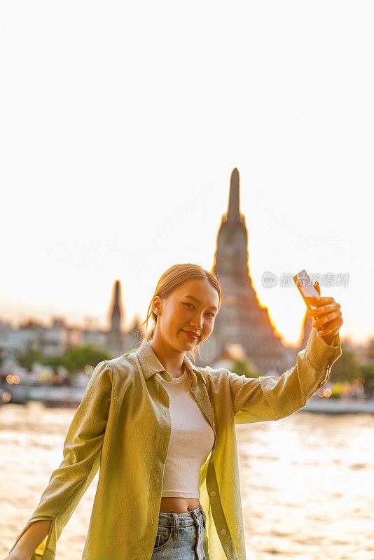 年轻的亚洲女子在泰国曼谷河边的阿龙寺的背景下捕捉自己日落的时刻