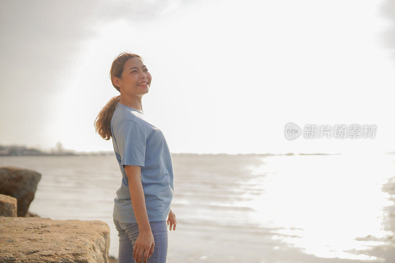 年轻的亚洲女子站在海滩的岩石上，带着幸福和希望的微笑望着远方。