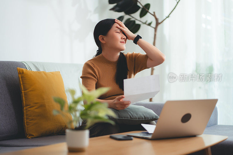 亚洲女性坐在沙发上，担心银行票据、抵押贷款债务和生活成本的账单支付问题。