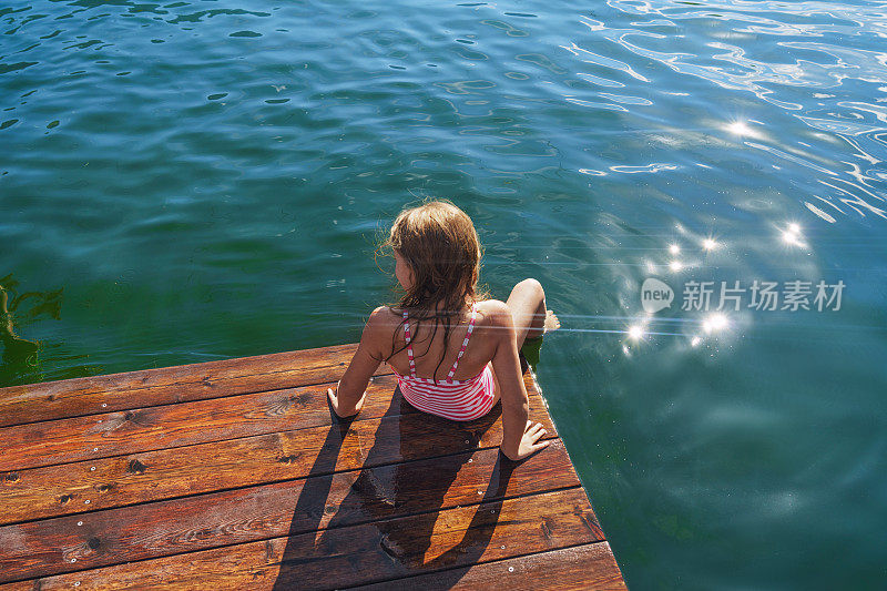 女孩坐在木制防波堤上，脚浸在水里。好好享受暑假。