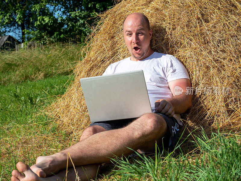 一位身穿t恤和短裤的中年男子正躺在黄色的干草堆上，手里拿着笔记本电脑。远程工作，培训。农村地区。男人脸上露出惊讶的表情，情绪激动