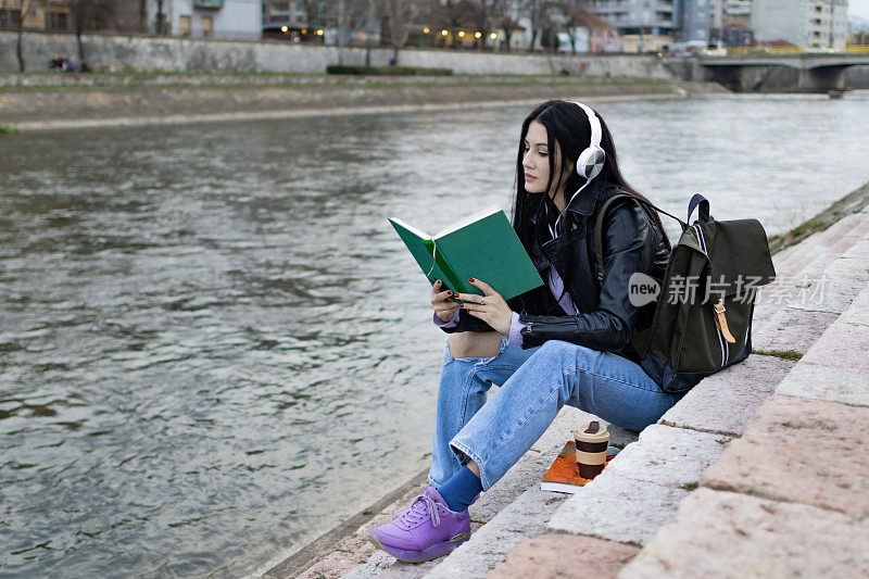 年轻漂亮的女大学生背着背包在河边玩。她戴着耳机，穿着皮夹克，等待着大学课程的开始。