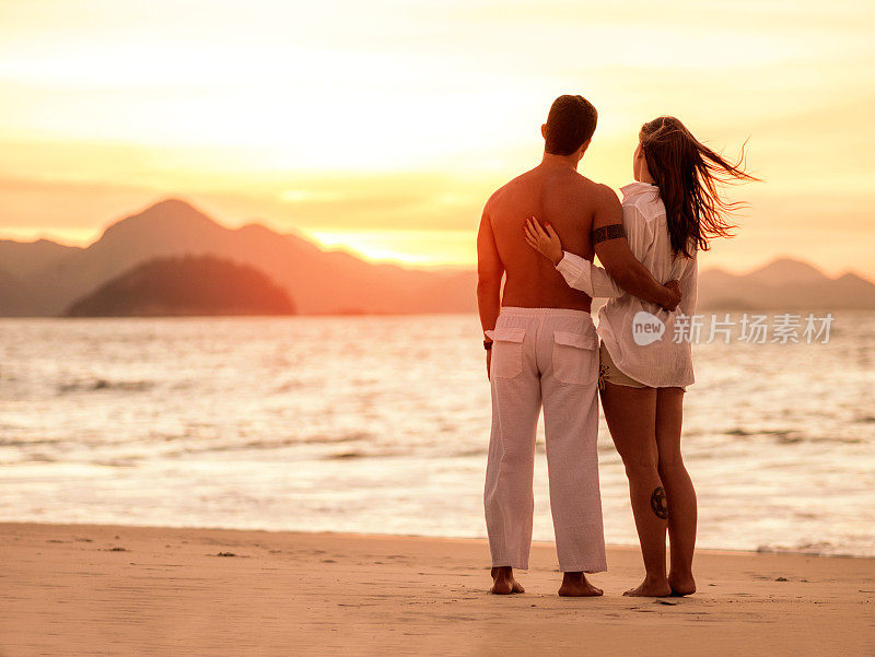在日落的海滩上拥抱的夫妇的后视图。