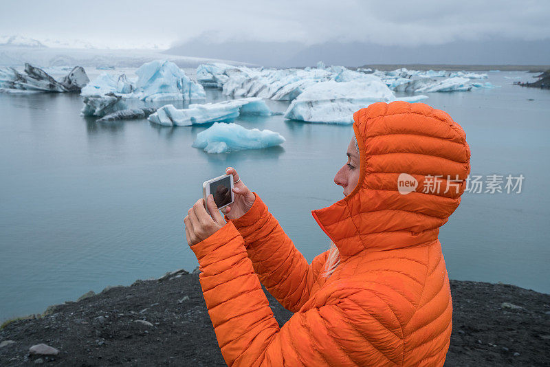 年轻女子用手机拍摄冰川泻湖，冰山漂浮在水面上