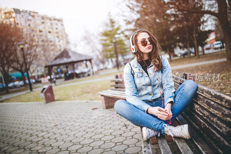 女人坐在公园的长椅上，戴着耳机听音乐。