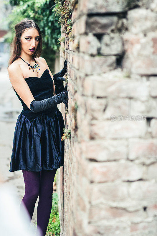 一个穿着黑裙子戴着手套的漂亮女孩站在一堵砖墙旁边