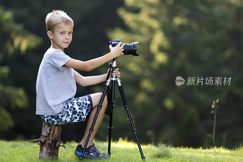 金发小男孩坐在树桩上的草地上用三脚架相机拍照。