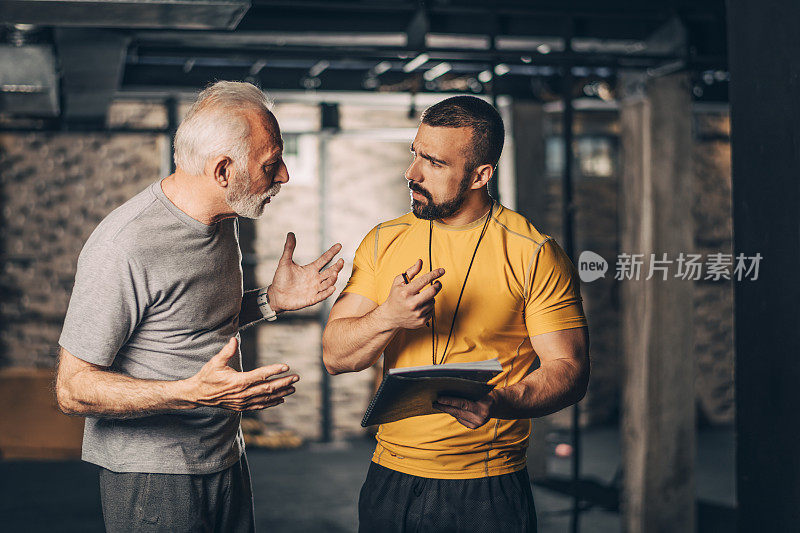 活跃的老年男性与他的个人健身教练谈话时，在一个交叉训练部分的健身房