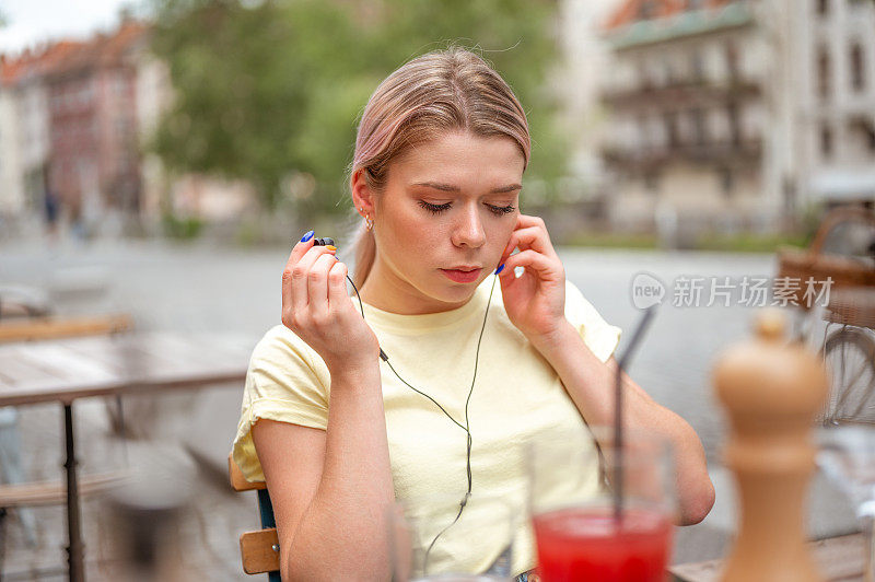 年轻的成年妇女坐在户外的咖啡馆里，使用手机和耳机