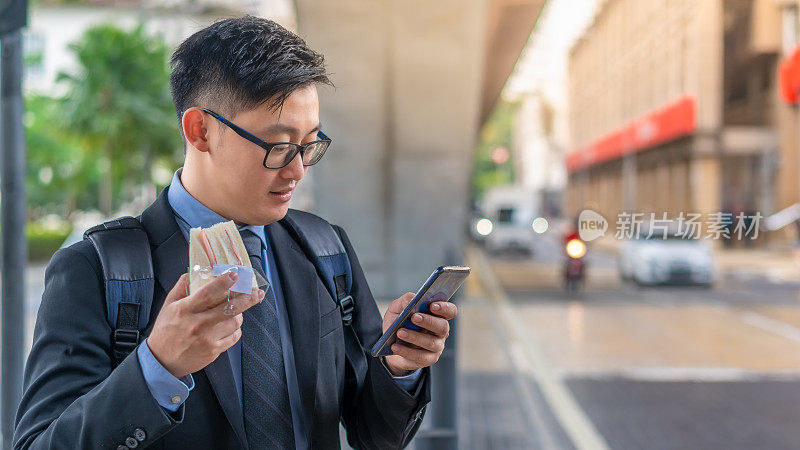 亚洲商人拿着午餐三明治，在城市街道上使用手机