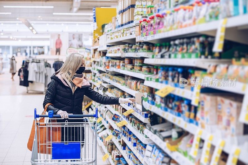 戴着防护口罩和橡胶手套的妇女站在超市里，推着购物车看着产品。