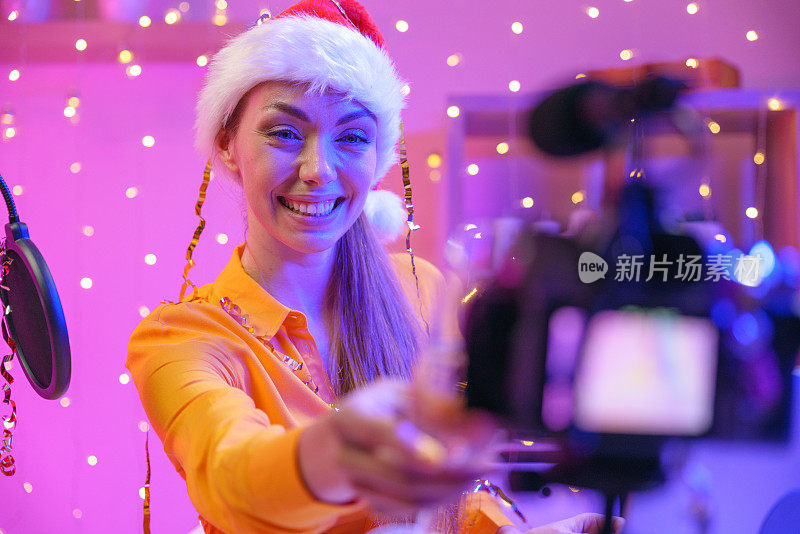 一个戴着圣诞老人帽子的视频博主女孩对着摄像机轻轻地摇着她的香槟。