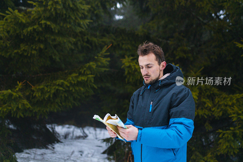 年轻的男性徒步旅行者穿着蓝色夹克，在雾蒙蒙的雪山中迷失了方向，看着地图