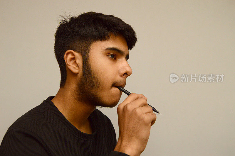 一个留着胡子的年轻的印度亚裔青年，手里拿着一支笔，嘴里衔着笔，陷入了沉思，在灰色的背景上看着空白，思考着，或打算做一个决定