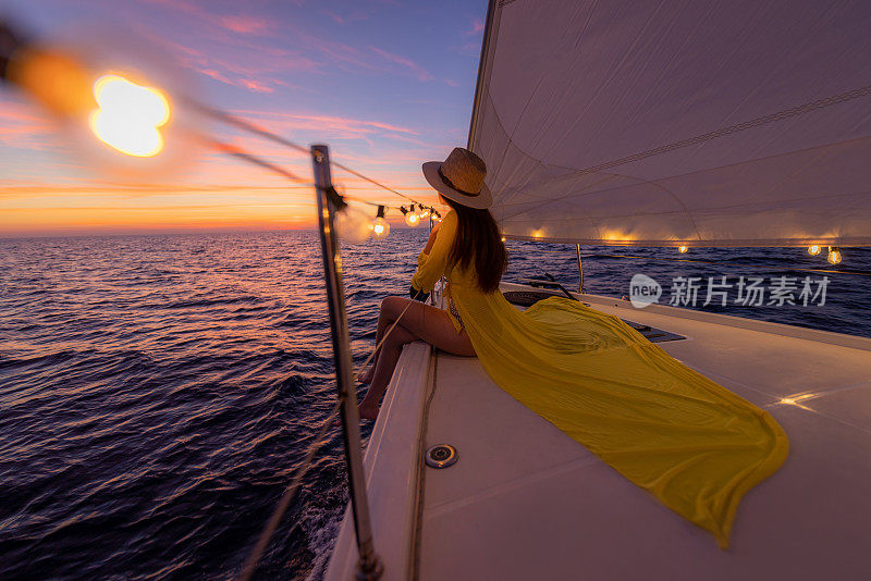戴着棕色长发、戴着帽子、穿着黄色连衣裙的女人坐在帆船的前甲板上，看着大海