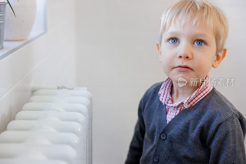 金发小男孩独自站在家里的暖气片旁，看起来有点激动