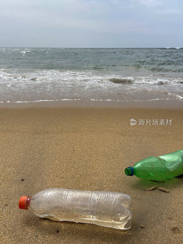 两个塑料水瓶在退潮时被冲到沙滩上的特写图像，海岸上的海洋垃圾和污染，乱扔的沙子，肮脏的海滩，关注前景