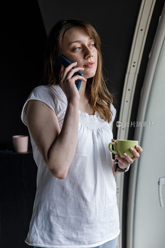 一位年轻女子站在窗边，一边喝着咖啡，一边讲着电话