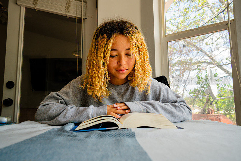 一个十几岁的非洲裔美国女孩，齐肩的头发遮住了脸，她穿着舒适的灰色运动衫，坐在餐桌旁，全神贯注地看书，钻研着迷人的书页。