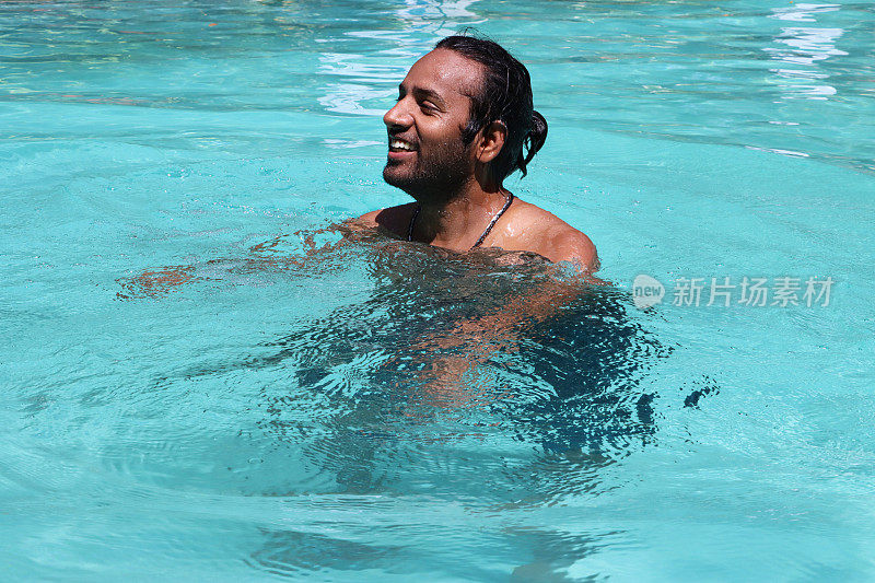 印度男子站在肩部深的清澈的水中，室外，游泳池，绿松石蓝色马赛克瓷砖被波纹水扭曲，游泳和飞溅，波涛汹涌的水面反射，重点在前景