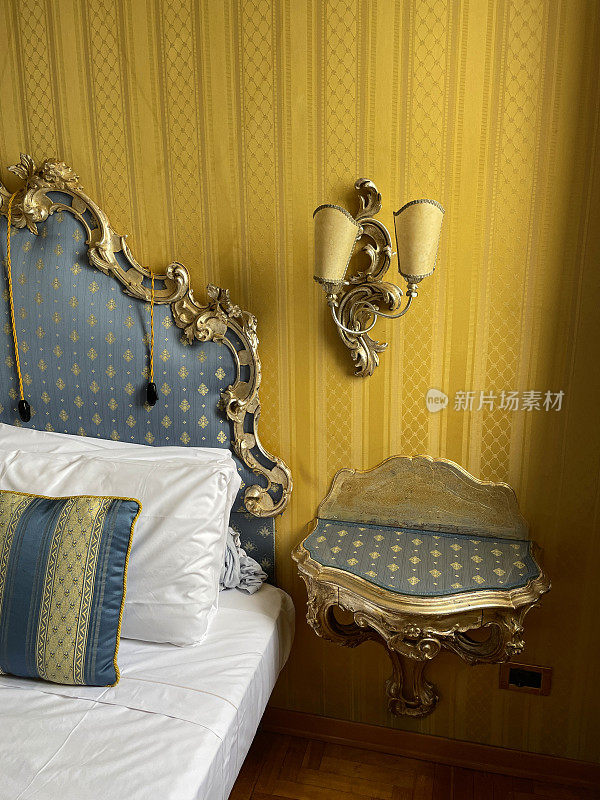 酒店房间双人床的图像，白色酒店枕头叠靠在华丽的框架，图案，软垫床头板，白色羽绒被套，蓝色和金色条纹靠垫，壁挂式电灯，床头柜，重点在前景