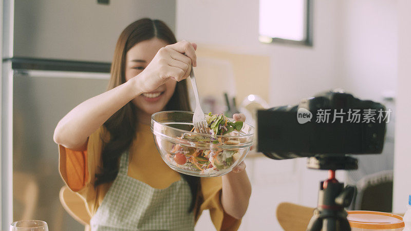 年轻微笑的美女网红美食博主在家里的厨房里吃东西和拍摄视频。社交媒体影响者概念。