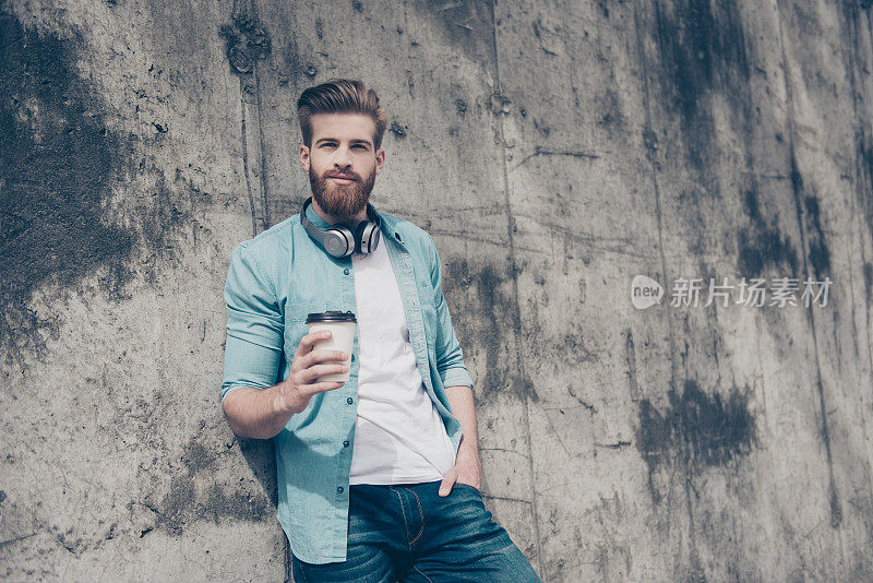 快乐的年轻时尚的大胡子男人，穿着牛仔裤衬衫，脖子上挂着耳机，靠在水泥墙上，喝着新鲜的咖啡