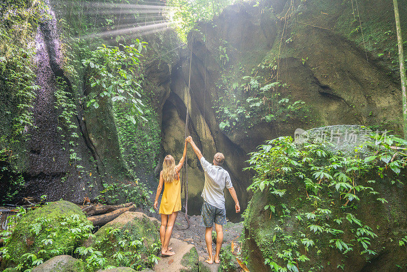 一对年轻夫妇站在热带雨林洞穴里，仰望着从岩石上射下来的壮丽阳光。人们旅行的奇妙欲望自然的概念