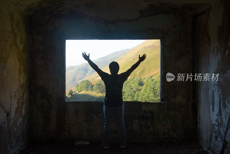 一个年轻人高举着手臂，站在一座废弃建筑的窗户前，看着美丽的山景