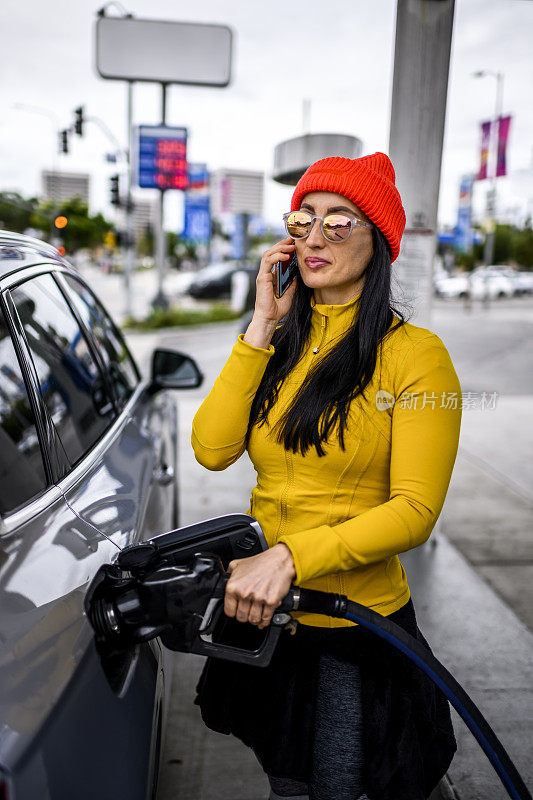 一位时髦的女士在加油站给她的车加油时，正在用智能手机打电话