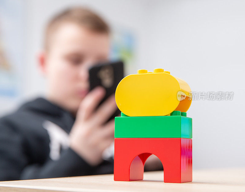 三个玩具积木站在桌子上与无聊的孩子使用他的手机在背景-库存照片