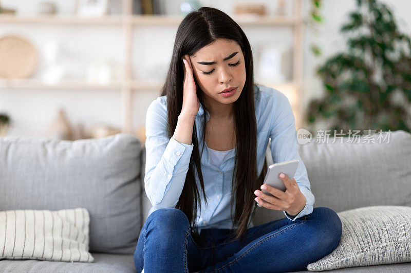 网络欺凌的概念。悲伤的韩国女孩坐在家里的沙发上拿着智能手机