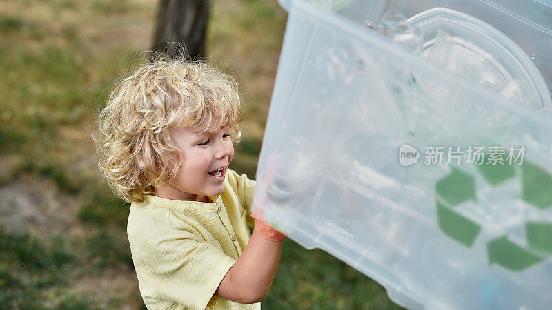 回收的概念。可爱的小男孩戴着橡胶手套拿着回收箱，微笑着在森林或公园收集塑料垃圾